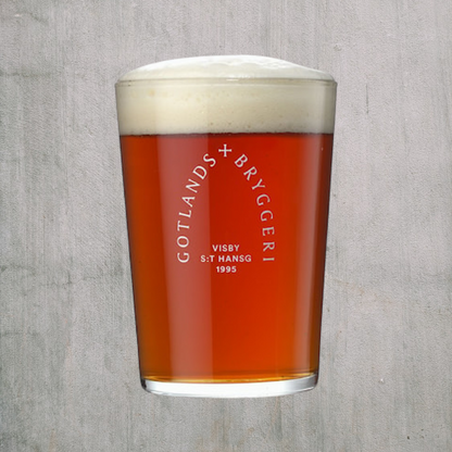 Glas med gotlands bryggeri logo öl i med skum på toppen mot grå bakgrund