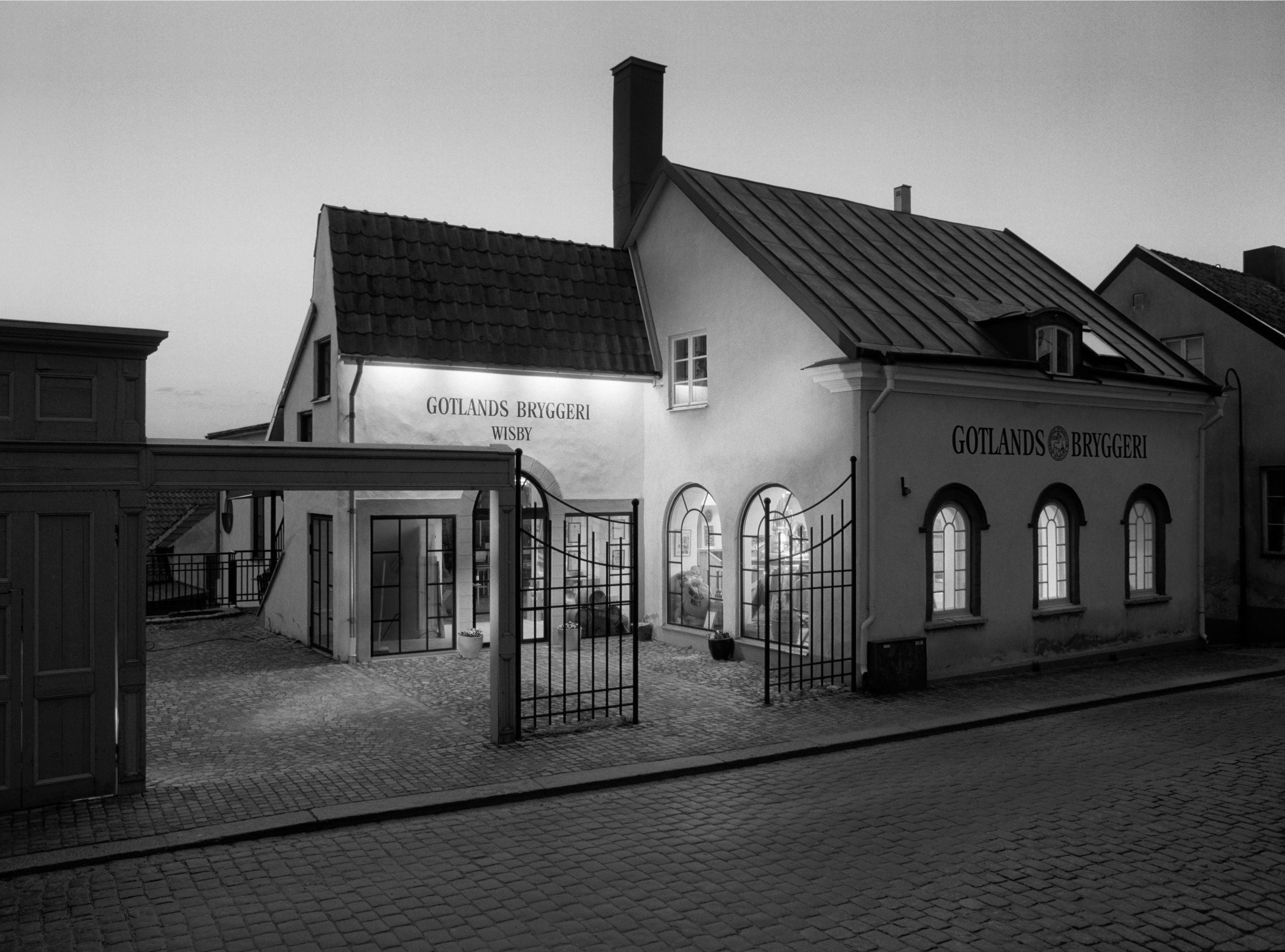 Gotlands Bryggerier byggnad upplyst av lampor inifrån och utifrån med kullerstensgata framför svart vit bild