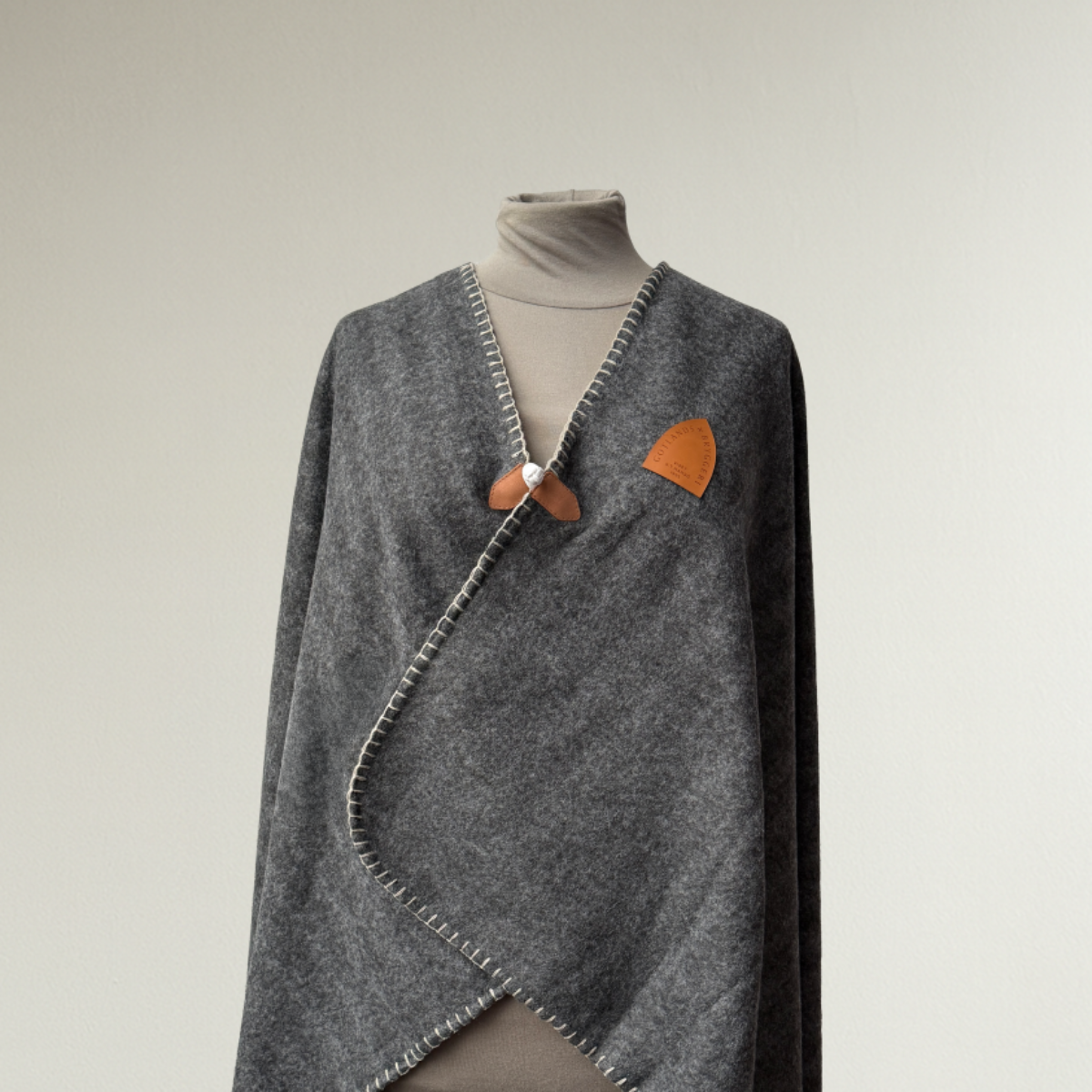 Grå pläd använd som poncho. Med brunt konstläder märke med logotyp. 